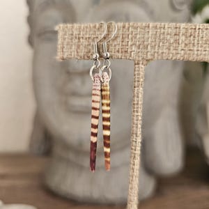 Pode incluir: Um par de brincos pendurados com um design natural listrado em tons de marrom, bege e roxo. Os brincos são suspensos por ganchos de orelha prateados. Os brincos são exibidos em um suporte de tecido bege.