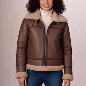 Puede incluir: Chaqueta marrón de piel sintética con cuello y ribetes de borreguillo beige. La chaqueta tiene cremallera, dos bolsillos delanteros y detalles de hebilla en el cuello. La chaqueta se lleva sobre un cuello alto blanco.