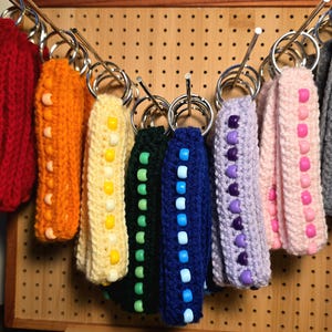 May include: A collection of crochet wristlet keychains in various solid colors including red, orange, yellow, dark green, blue, lavender, pink, and gray. Each keychain features a row of matching plastic beads and a silver metal split ring.