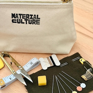 May include: A beige canvas pouch with the text "MATERIAL CULTURE" in black, a gold zip, and sewing supplies. The supplies include scissors, a tape measure, needles, pins, thread, and a bar of yellow wax.
