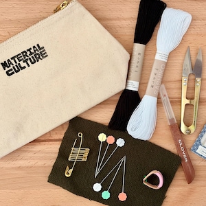 以下が含まれることがあります： 「MATERIAL CULTURE」の文字が入ったベージュ色のキャンバス地のポーチ、黒と白の刺繍糸、金色のハサミ、ピン、小さなカッター、針のパッケージなど、縫製と刺繍用品のコレクション。