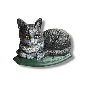 May include: A decorative cat figurine in a resting pose. The cat is primarily black with gray and white accents, yellow eyes, and a pink nose. It rests on a green, leaf-shaped base. The figurine is likely made of resin or a similar material.