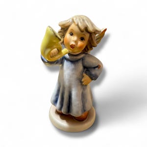 Op de afbeelding: Een keramieken beeldje van een engel die op een gele hoorn blaast. De engel heeft blond haar, roze wangen en draagt een blauw-witte jurk. Het beeldje staat op een ronde, gebroken witte basis.