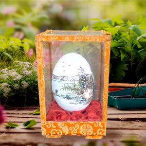 Puede incluir: Un huevo blanco con una escena de paisaje pintada dentro de una vitrina de vidrio transparente con una base de terciopelo rojo y un exterior de terciopelo dorado.