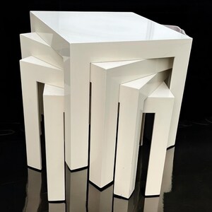 May include: A set of four white nesting tables with a glossy finish. The tables are square and have a geometric design, with each table slightly smaller than the one beneath it. The tables are stacked together, creating a tiered effect.