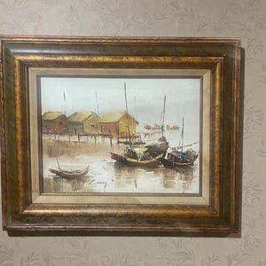 May include: Oil painting of a coastal scene with boats and huts on stilts. The artwork features a wooden frame with gold accents. The boats are in the water, and the huts are in the background. The painting has a muted color palette.
