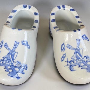 May include: Two white ceramic clogs featuring blue windmill and landscape patterns. The clogs have a glossy finish and blue detailing around the opening. A decorative item, reminiscent of Dutch design.