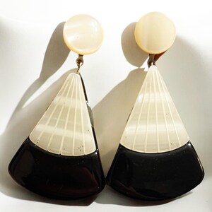 May include: A pair of vintage clip-on earrings. Each earring features a cream-colored circular top and a geometric, fan-shaped pendant. The pendant is divided into a black, glossy bottom and a cream-colored, vertically striped top section.