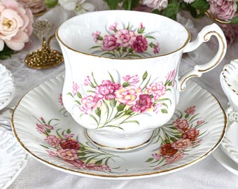 May include: A white teacup and saucer set with a floral pattern of pink and yellow carnations. The set has a gold trim around the edges.