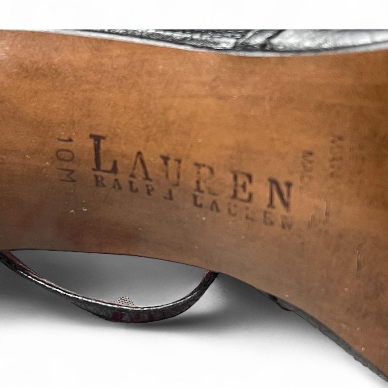 May include: Close-up of the sole of a black high-heeled shoe. The brand name "LAUREN RALPH LAUREN" is stamped in dark ink on the brown sole. The shoe size is 10M.