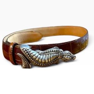May include: A brown belt with a textured, embossed surface and a polished silver-tone metal buckle shaped like a stylized alligator or crocodile. The belt features a smooth, light tan interior lining and a dark brown exterior finish.