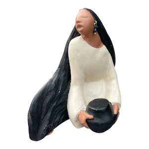 Könnte beinhalten: Handgefertigte Figur einer Person in einem weißen Kleid mit langen schwarzen Haaren. Die Figur hält einen schwarzen Hut. Die Person trägt Ohrringe mit blauen und goldenen Akzenten. Die Figur ist ein einzigartiges Kunstwerk.