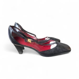May include: Black leather heeled shoes with a square toe and a strap across the foot. The shoes have a red interior and a gold buckle. The heels are approximately 3 inches high.