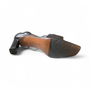 May include: Black leather heeled shoe with a square toe and a thick, block heel. The sole is brown with the word "LAUREN" stamped on it. The shoe has a strap that wraps around the ankle.