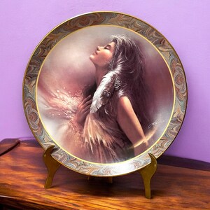 May include: A decorative porcelain plate with a gold rim features a painting of a woman with long dark hair, wearing a feathered headdress, with her eyes closed and head tilted back. The plate is on a wooden stand.