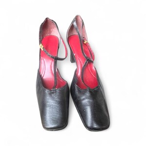 May include: A pair of black, textured leather heels with a square toe and a T-strap design. The interior is a vibrant red, and the heels have a small gold buckle. The shoes are set against a white background.