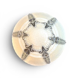 May include: A round, off-white ceiling light fixture with a textured surface. It features silver-colored leaf and scroll accents around the perimeter. The center has a floral design.