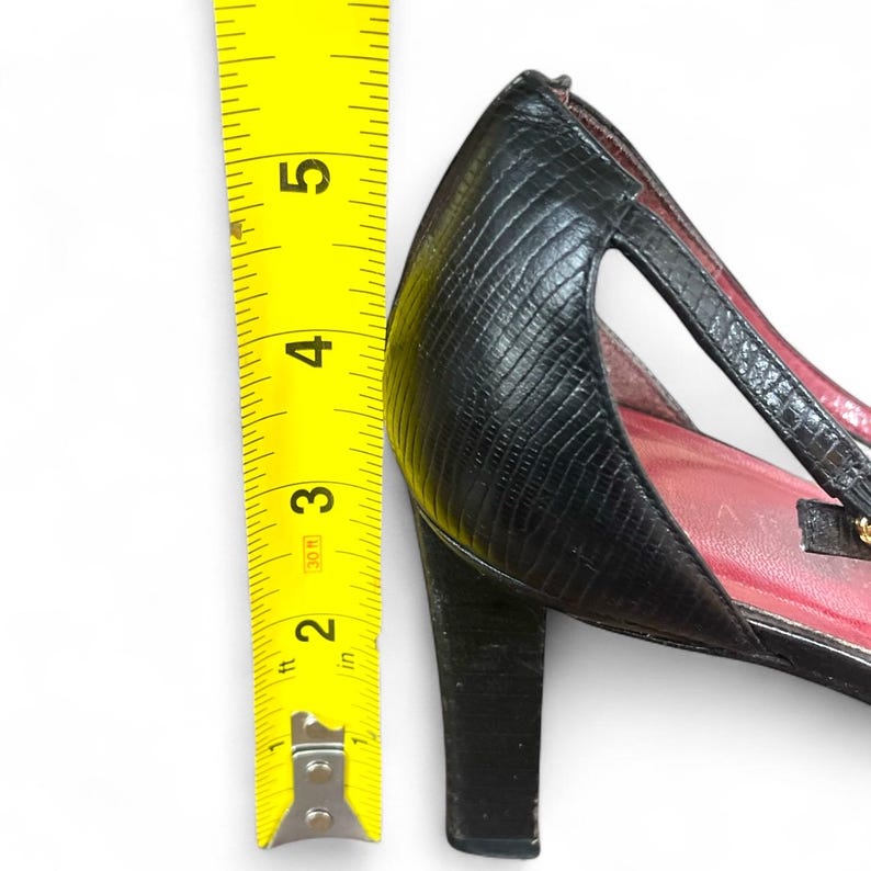 May include: Black high-heeled shoe with a textured pattern. The heel measures approximately 3 inches. A yellow measuring tape is positioned next to the shoe, showing the height of the heel.