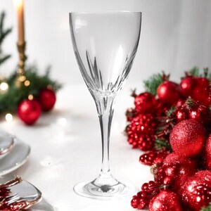 Może przedstawiać: Szklanka do wina z przezroczystego szkła z delikatną n&oacute;żką i szeroką czaszą. Szklanka stoi na białym obrusie z czerwonymi i zielonymi ozdobami świątecznymi.