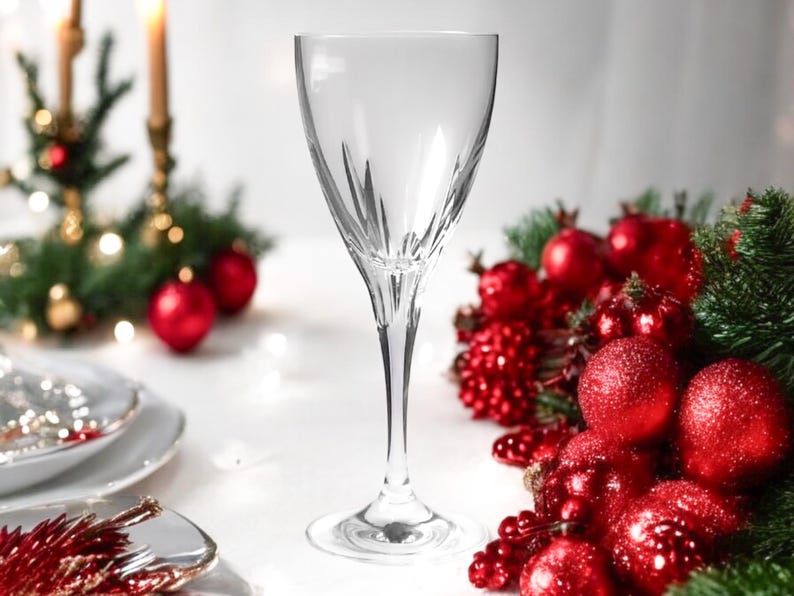Może przedstawiać: Szklanka do wina z przezroczystego szkła z delikatną n&oacute;żką i szeroką czaszą. Szklanka stoi na białym obrusie z czerwonymi i zielonymi ozdobami świątecznymi.
