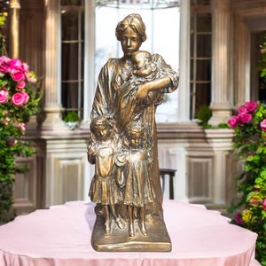 May include: A bronze sculpture of a woman holding a child in her arms and two other children by her side. The sculpture is on a pedestal and is surrounded by pink flowers.