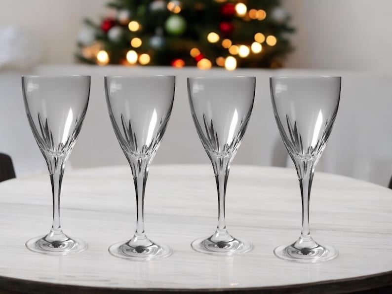 Może przedstawiać: Cztery przezroczyste szklane kieliszki do wina z ozdobnym wzorem n&oacute;żki. Kieliszki są ustawione w rzędzie na białym stole.