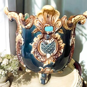 May include: A large, ornate, blue and brown ceramic planter with gold accents and three legs. The planter has a decorative design with a turquoise center piece.