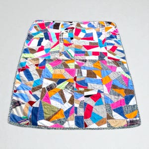 Op de afbeelding: Een kleurrijke patchworkquilt met een verscheidenheid aan stofpatronen en kleuren, waaronder roze, blauw, oranje en bruin. De quilt is rechthoekig met een gedetailleerde rand. De quilt is gemaakt van vele kleine stukjes stof die aan elkaar zijn genaaid.