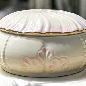 May include: A ceramic trinket box with a domed lid. The lid is white with a pink edge and gold trim. The body is light gray with pink floral accents and a beaded design. A decorative storage container.