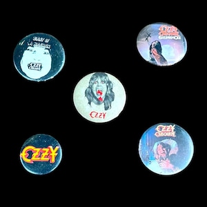 May include: A collection of five round metal pinback buttons featuring images of Ozzy Osbourne. The buttons display album art and portraits, with text including "Ozzy" and "Diary of a Madman."