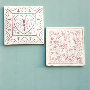 Puede incluir: Dos baldosas cuadradas blancas con acabado envejecido. Una baldosa presenta un diseño de corazón con flechas, la otra un patrón floral. Ambas baldosas tienen un diseño rojo rosado y están sobre un fondo turquesa.