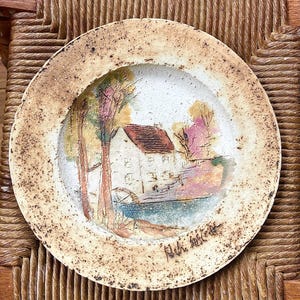 May include: Round ceramic plate with a hand-painted landscape scene. The plate features a white building with a red roof, trees, and a body of water. The rim has a speckled, textured appearance with a light brown color.