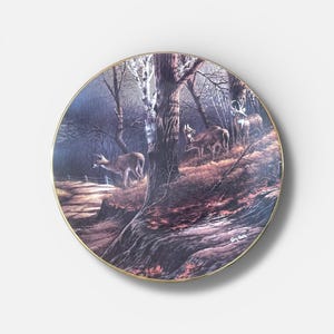 May include: A decorative plate featuring a forest scene with deer. The plate has a gold rim and a detailed painting of deer in a wooded area with trees and a path. The artwork is signed by the artist.