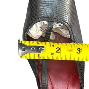 May include: Black leather sandal with a T-strap design. The shoe has a red insole and a textured exterior. A measuring tape shows the shoe's width is approximately 2 inches.