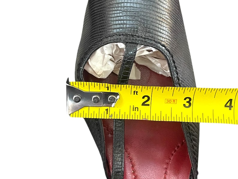 May include: Black leather sandal with a T-strap design. The shoe has a red insole and a textured exterior. A measuring tape shows the shoe's width is approximately 2 inches.