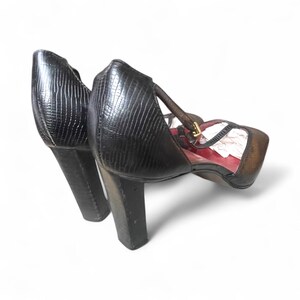 May include: A pair of black heeled sandals with a textured, reptile-like pattern. The sandals have a T-strap design with a gold-colored buckle and a square toe. The heels are approximately 3 inches tall.