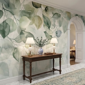 May include: A hallway with a watercolor-style mural of green eucalyptus leaves. A dark wood console table holds two white lamps with beige shades, a blue and white vase, and books. A patterned rug runs along the floor.