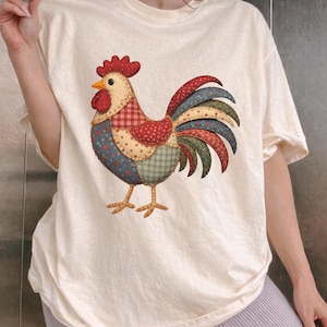 Peut inclure: T-shirt crème avec un motif de coq coloré. Le coq est fait de patchwork avec des motifs rouges, bleus, verts et jaunes. Le t-shirt a des manches courtes et une coupe décontractée, idéal pour un usage quotidien.