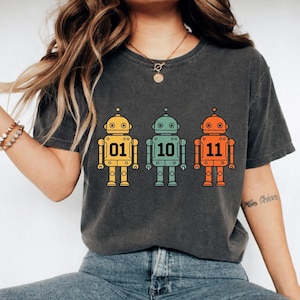 Peut inclure: T-shirt gris foncé avec trois illustrations de robots colorés. Chaque robot est jaune, turquoise et orange, avec les chiffres 01, 10 et 11. Ils ont des yeux circulaires et des antennes. Vêtement décontracté.