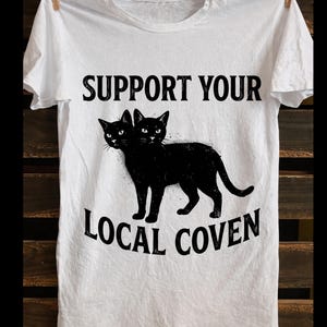 Peut inclure: T-shirt blanc avec l'inscription noire "SUPPORT YOUR LOCAL COVEN", au-dessus et en dessous d'une illustration de chat noir à deux têtes. Le t-shirt est suspendu à une corde à linge sur un fond en bois.