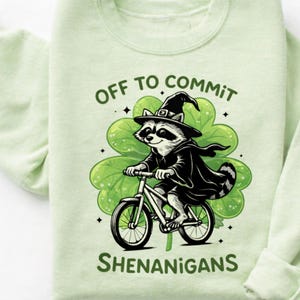 Peut inclure: Sweat-shirt vert clair avec un dessin d'un raton laveur à vélo, portant un chapeau de sorcière et une cape, devant un trèfle vert. Le texte dit "OFF TO COMMIT SHENANIGANS".