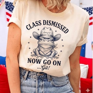 Puede incluir: Camiseta color crema con una rana de dibujos animados con sombrero de vaquero y pañuelo. El texto "CLASS DISMISSED NOW GO ON ...Git!" está impreso encima y debajo de la ilustración de la rana. El sombrero tiene el texto "Love y'all".