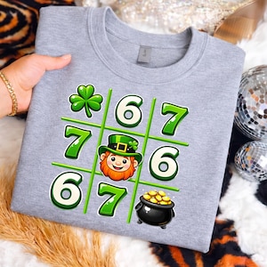 Peut inclure: Un sweat-shirt gris avec un motif de morpion sur le thème de la Saint-Patrick. Le motif comprend un trèfle vert, les chiffres 6 et 7, un lutin et un pot d'or. Le sweat-shirt est tenu par une personne.