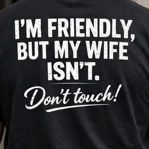 May include: Black t-shirt with white text. The text reads "I'M FRIENDLY, BUT MY WIFE ISN'T. Don't touch!" in a bold, sans-serif font, with the last line in a cursive font. The shirt is a casual wear item.