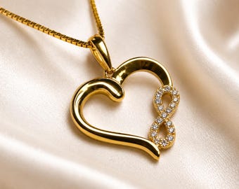 Puede incluir: Un collar de tono dorado con un colgante en forma de corazón. El colgante presenta un acabado dorado liso y pulido en un lado, y un símbolo de infinito incrustado con pequeñas piedras claras y brillantes en el otro, suspendido de una cadena.