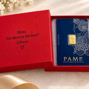 Puede incluir: Una peque&ntilde;a barra de oro en una tarjeta de ensayo azul de PAMP Suisse, presentada dentro de una caja de regalo roja. El interior de la tapa de la caja tiene el texto impreso en negro: Mom, You deserve the best! Johnny, seguido de un coraz&oacute;n.