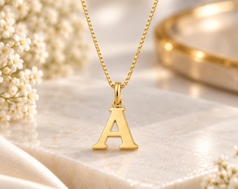 Puede incluir: Un collar de tono dorado con un colgante pulido en forma de letra A suspendido de una delicada cadena de eslabones cuadrados. El colgante está unido a la cadena mediante una pequeña anilla circular, con un acabado metálico liso y brillante.