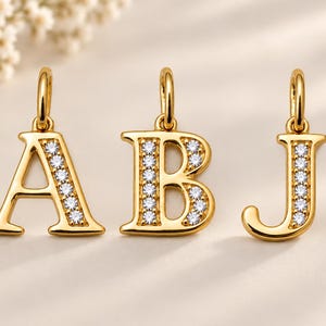 May include: Three gold letter pendants featuring the letters A, B, and J. Each pendant has a polished gold finish and is accented with a vertical row of small, clear, round-cut stones embedded along the front face of the letter characters.