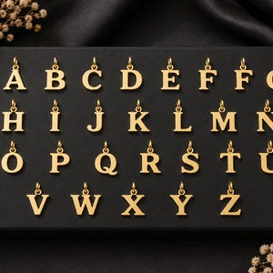 Könnte beinhalten: Eine Sammlung goldfarbener Metallanhänger in Buchstabenform, die in alphabetischer Reihenfolge auf einer schwarzen rechteckigen Anzeigetafel angeordnet sind. Jeder einzelne Buchstabenanhänger hat oben eine kleine Öse zur Befestigung.