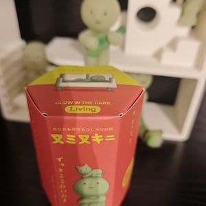 May include: A red box with the text "GLOW IN THE DARK Living" and Japanese characters. The box features a green cartoon figure with a cat on its head. A green figure playing a guitar is in the background.